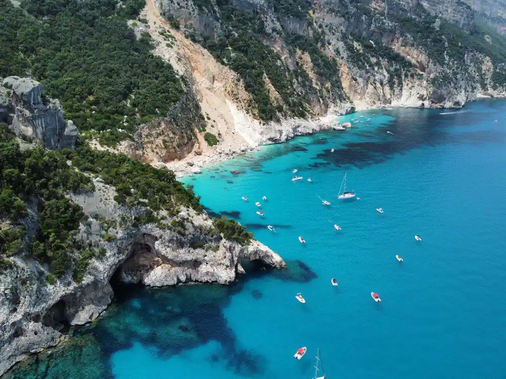 Aerial view of turquoise bay with multiple anchored sailboats and cliffs