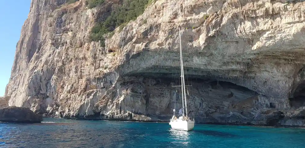 White sailboat anchored alone under large natural cave in limestone cliff